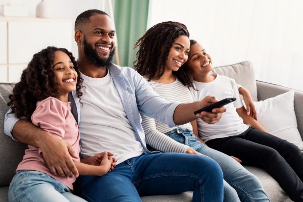 Comfort And Relax. Portrait of happy loving black family of four people watching tv, smiling parents embracing their two children sitting on couch in the living room, dad holding remote control